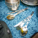 Image of GSI stainless steel 3-piece camping cutlery sets with spoon, fork, and serrated knife laid out on blue patterned camp table beside metal cookware, angled top view.
