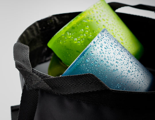 Close-up image of a black washbasin bag containing a wet blue and a wet green cup with water droplets, against a neutral background.