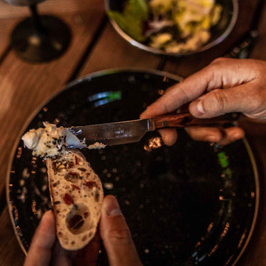 Image of stainless steel serrated table knife with wooden handle spreading soft cheese onto sliced bread, close-up dining scene with plate and salad in background.