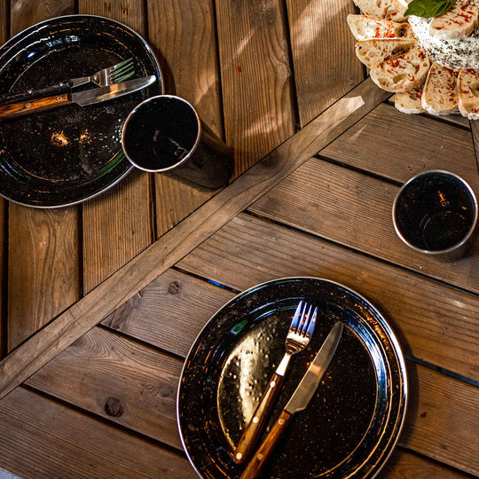Image of stainless steel table knife and fork with wooden handles placed on black enamel plates, top-down dining table view with bread and cups on rustic wood surface.