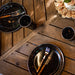 Image of stainless steel table knife and fork with wooden handles placed on black enamel plates, top-down dining table view with bread and cups on rustic wood surface.