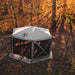 Image of a fully deployed beige Gazelle screen tent with black mesh walls, set up on a leaf-covered forest floor. The photo is taken from a slightly elevated angle, with sunlight filtering through the yellow and orange autumn trees in the background