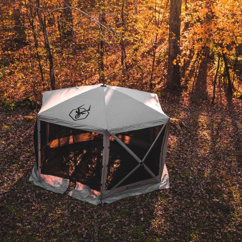 Image of a fully deployed beige Gazelle screen tent with black mesh walls, set up on a leaf-covered forest floor. The photo is taken from a slightly elevated angle, with sunlight filtering through the yellow and orange autumn trees in the background