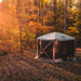 Image of a fully set up beige Gazelle screen tent with black mesh walls, placed on a forest floor covered in autumn leaves. The image is a wide-angled shot, with sunbeams filtering through the yellow, orange, and green trees in the background