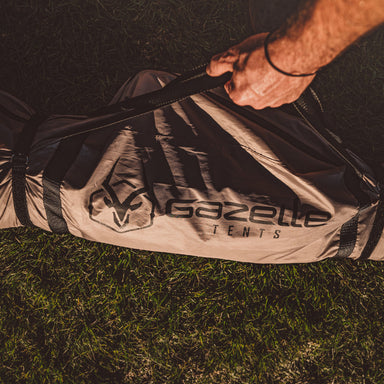 Image of a person's hand grabbing the black carrying strap of a folded, beige Gazelle tent bag with a black logo, lying on a patch of green grass