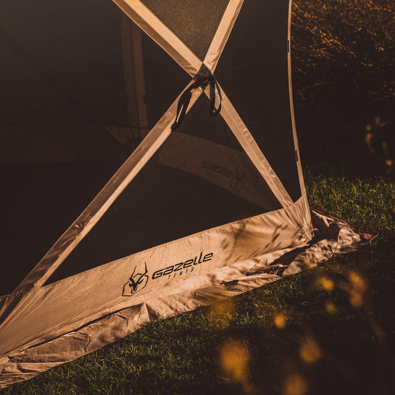 Image of the lower corner of a beige Gazelle screen tent with black mesh walls, set up on green grass. The tent's "Gazelle Tents" logo is visible on the beige fabric skirt