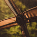 Image of the central hub and frame of a beige gazebo tent, showing the connections of the metal poles and the fabric panels. The image is a close-up, with a blurred, green background seen through the tent's black mesh
