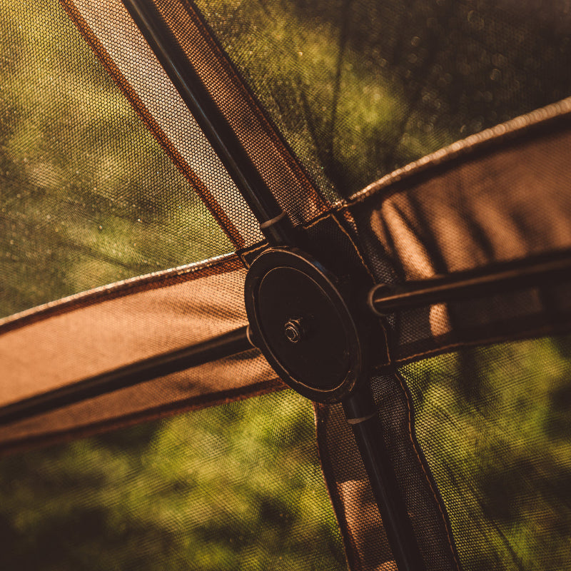 Image of the central hub and frame of a beige gazebo tent, showing the connections of the metal poles and the fabric panels. The image is a close-up, with a blurred, green background seen through the tent's black mesh