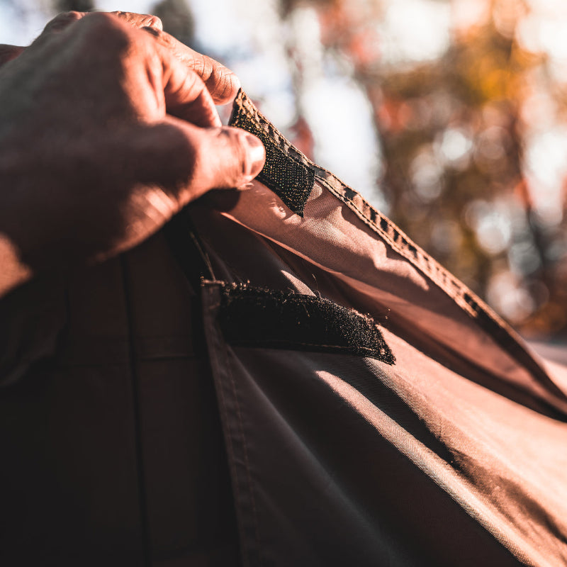 Image of a person's hand touching the black hook and loop fastener (velcro) on the corner of a beige gazebo tent, with a blurry background of autumn trees. The image highlights the attachment system of the tent's fabric panels