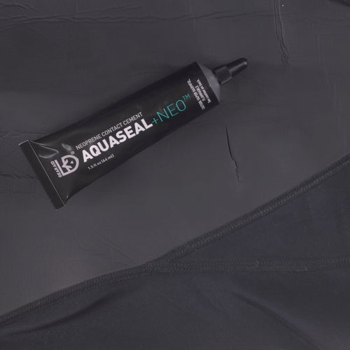 Image of Gear Aid Aquaseal Neo contact cement tube placed on dark neoprene fabric with visible seam and tear, angled top view on a dark background.