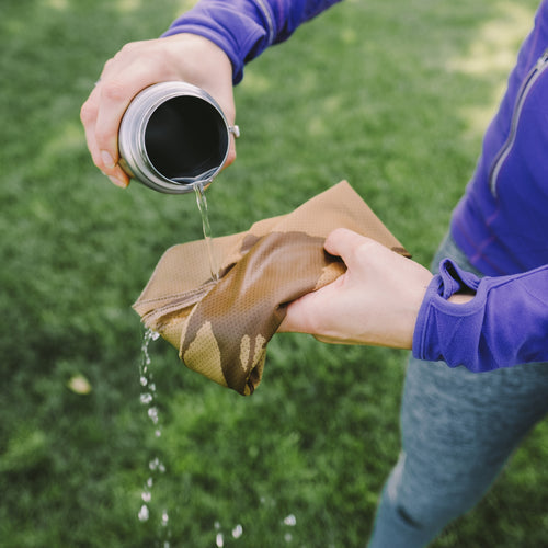 Image of Gear Aid cooling towel being activated with water poured from a bottle, tan fabric held in hands outdoors, close-up action view on green grass background.