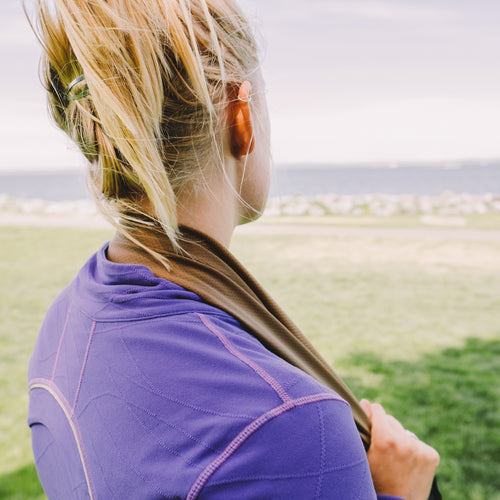 Image of person wearing a Gear Aid cooling towel draped around the neck, tan fabric in use outdoors near grass and water, rear angled lifestyle view.