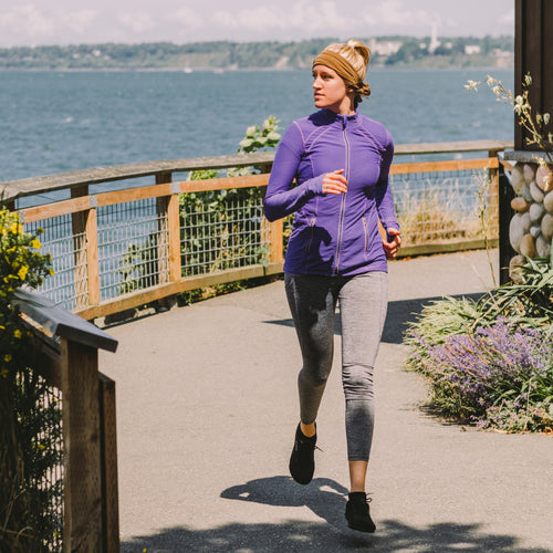 Image of person jogging outdoors wearing a Gear Aid cooling towel around the head, tan fabric in active use on a waterfront path, full-body lifestyle view.