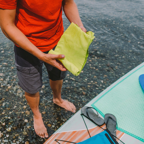 Image of person holding a Gear Aid microfiber towel near water and paddleboard, green fabric in use outdoors, top-down lifestyle view on rocky shoreline.