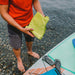 Image of person holding a Gear Aid microfiber towel near water and paddleboard, green fabric in use outdoors, top-down lifestyle view on rocky shoreline.