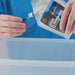 Image of Gear Aid Revivex Odor Eliminator being poured from a flexible pouch into a blue measuring cap, hands visible above a clear plastic bin, close-up angled view.