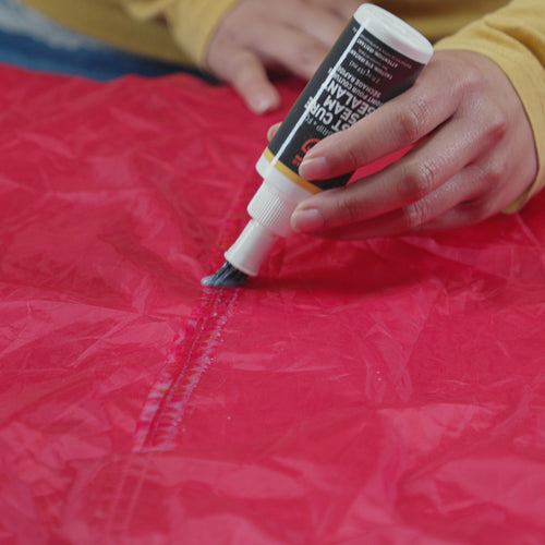 Image of Gear Aid Seam Grip FC Fast Cure Seam Sealant being applied with a brush along a tent seam on red fabric, close-up angled view showing waterproofing process.