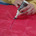 Image of Gear Aid Seam Grip FC Fast Cure Seam Sealant being applied with a brush along a tent seam on red fabric, close-up angled view showing waterproofing process.