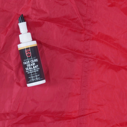 Image of Gear Aid Seam Grip FC Fast Cure Seam Sealant bottle placed on red tent fabric next to a sealed seam, top-down view showing waterproofing result.