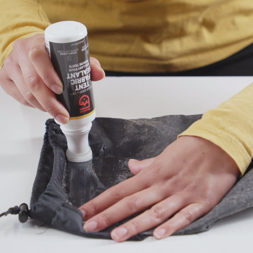 Image of Gear Aid Seam Grip TF Tent Fabric Sealant being applied to tent fabric with squeeze bottle, hands smoothing sealant on dark material, close-up angled view on neutral background.