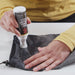 Image of Gear Aid Seam Grip TF Tent Fabric Sealant being applied to tent fabric with squeeze bottle, hands smoothing sealant on dark material, close-up angled view on neutral background.