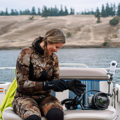 Image of woman in camo wetsuit sitting on boat bench adjusting black neoprene gloves beside underwater camera housing, lakeside background view.