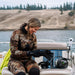 Image of woman in camo wetsuit sitting on boat bench adjusting black neoprene gloves beside underwater camera housing, lakeside background view.
