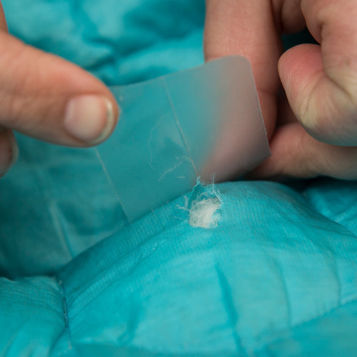 Image of hands applying a clear repair patch over a small tear in teal outdoor fabric, close-up top view showing patch alignment on textured material.