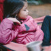 Image of a child wearing a pink insulated jacket with a black tree-shaped adhesive repair patch applied, seated outdoors in a camping chair with a casual side view.