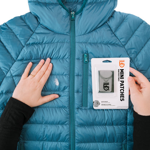 Image of Gear Aid Tenacious Tape Mini Patches demonstrated on a blue down jacket with hands smoothing fabric and retail packaging shown, front-facing repair example on white background.