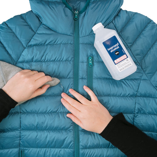 Image of blue down jacket being cleaned with cloth and isopropyl alcohol before repair, preparation step shown for applying Gear Aid Tenacious Tape Mini Patches on white background.