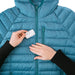Image of Gear Aid Tenacious Tape Mini Patch being positioned by hand over a tear on a blue down jacket, close-up repair step shown on white background.