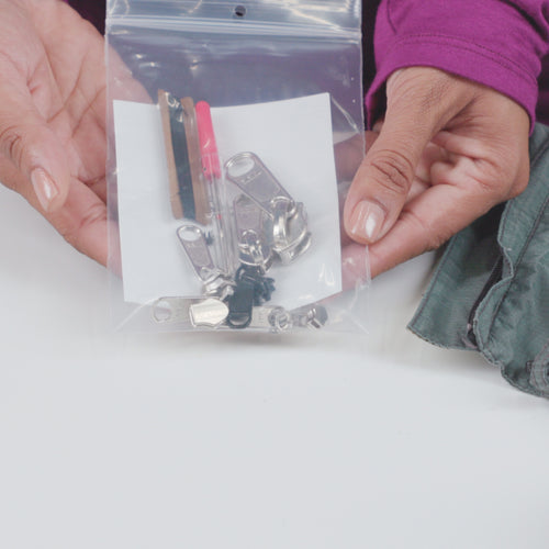 Image of hands holding a clear zip bag containing zipper repair kit components including metal sliders and needle tool, front view on white background.