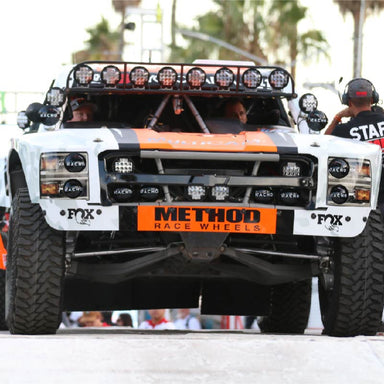 Front view of a white, orange, and black off-road racing truck with a row of circular lights mounted on its roof and two smaller, rectangular light bars mounted on the front bumper. The driver and passenger are visible inside the vehicle