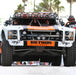 Front view of a white, orange, and black off-road racing truck with a row of circular lights mounted on its roof and two smaller, rectangular light bars mounted on the front bumper. The driver and passenger are visible inside the vehicle