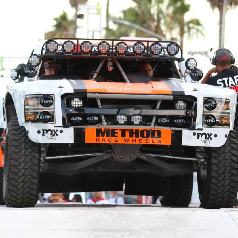Front view of a white, orange, and black off-road racing truck with a row of circular lights mounted on its roof and two smaller, rectangular light bars mounted on the front bumper. The driver and passenger are visible inside the vehicle