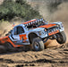 Image of a white and orange off-road racing truck with "FOX" and "METHOD RACE WHEELS" logos, kicking up dirt as it lands from a jump. The truck has a row of circular lights on its roof and is shown in action on a dirt track with trees in the background