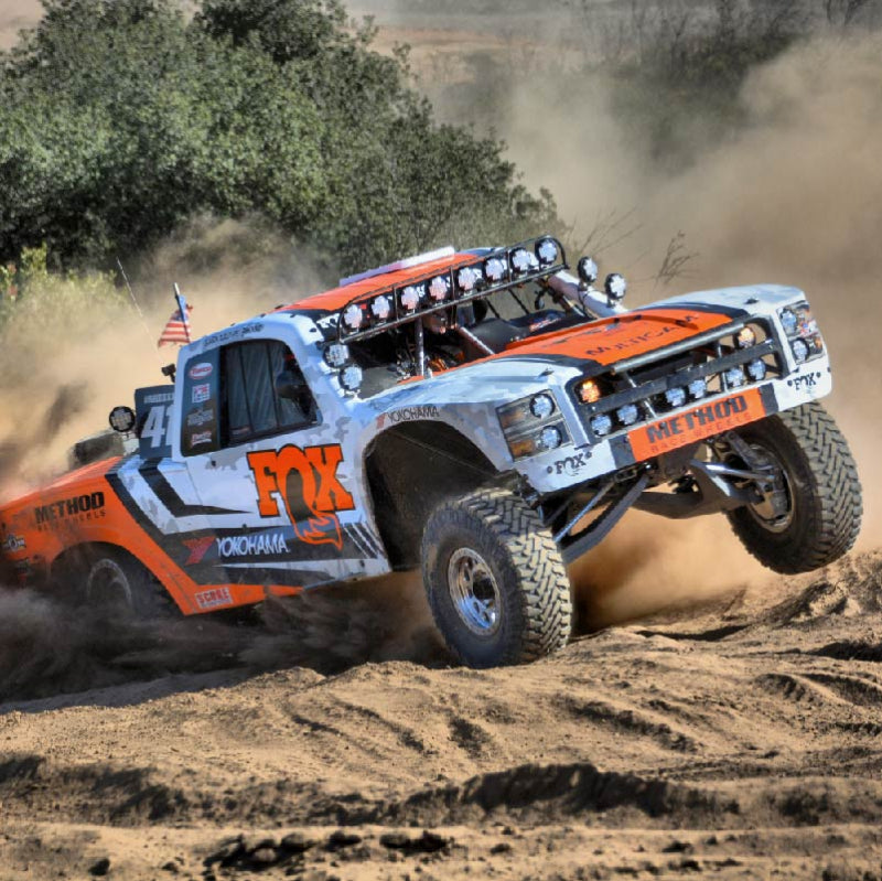 Image of a white and orange off-road racing truck with "FOX" and "METHOD RACE WHEELS" logos, kicking up dirt as it lands from a jump. The truck has a row of circular lights on its roof and is shown in action on a dirt track with trees in the background