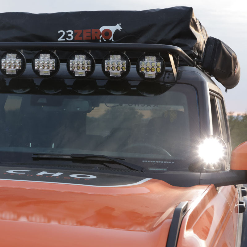 Angled front view of an orange and black off-road vehicle with a "23ZERO" roof top tent, a row of five round LED lights on the roof rack, and a single LED light mounted on the side mirror