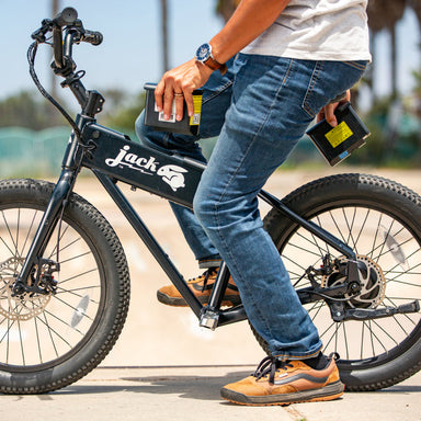 Image of a person sitting on a black JackRabbit fat-tire electric bike, holding an extra battery pack in their hands on a paved road.