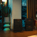 Image of Jackery home backup power system with main power station and stacked expansion batteries connected by cables, black units with orange logos placed indoors near a window, angled front view in home interior setting.