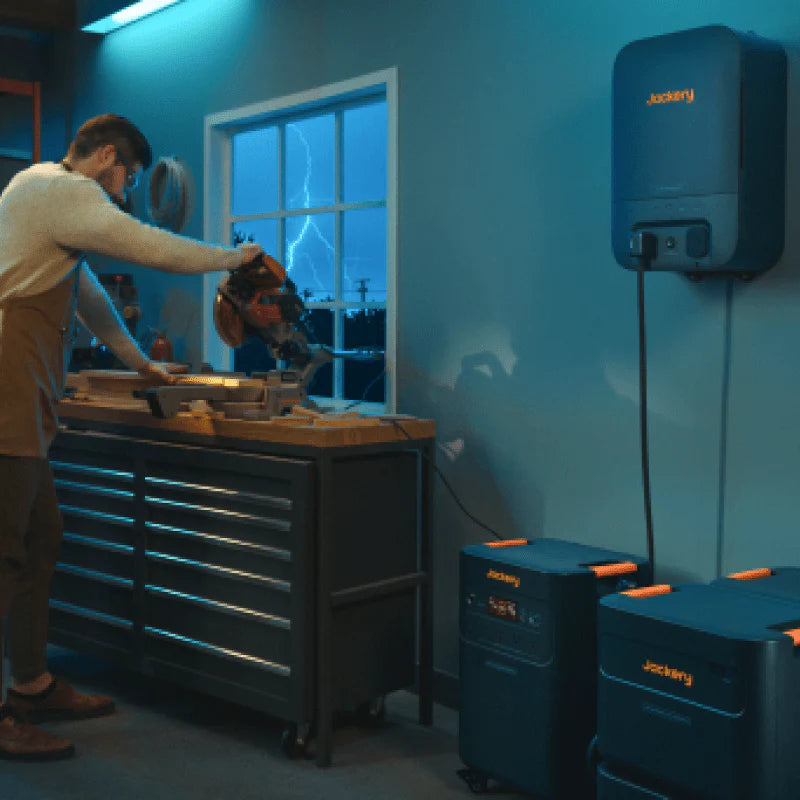 Image of Jackery home battery system with wall-mounted unit and expansion batteries powering workshop tools, black and orange units connected and in use, angled side view in indoor garage setting.