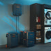 Image of Jackery home energy storage system with wall-mounted control unit, main power station, and stacked expansion batteries, black units with orange accents arranged in a laundry room, angled front view in indoor home setting.