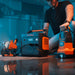 Image of Jackery Explorer 2000 portable power station powering work lights and equipment, black and orange unit with illuminated display in use beside tools, angled front view in indoor workshop setting.