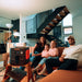 Image of Jackery Explorer 2000 portable power station powering a projector in a living room, black and orange unit with connected devices beside a family on a sofa, angled front view in indoor home setting.