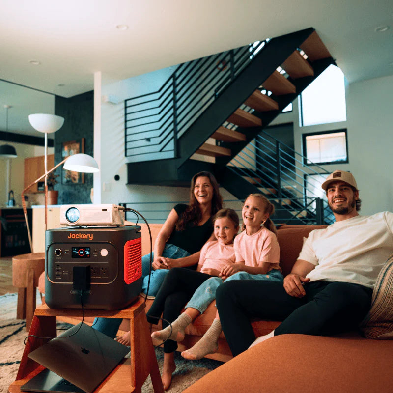 Image of Jackery Explorer 2000 portable power station powering a projector in a living room, black and orange unit with connected devices beside a family on a sofa, angled front view in indoor home setting.