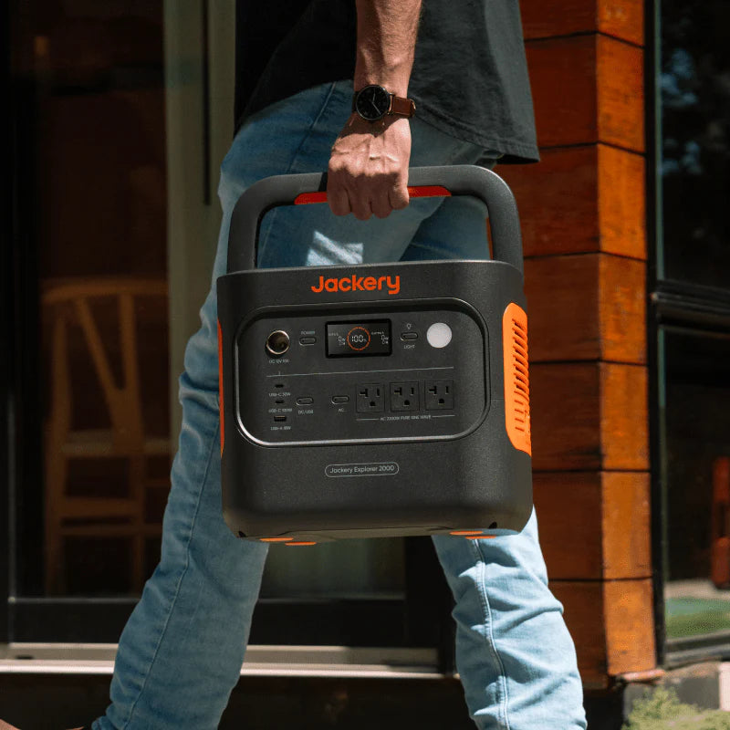 Image of Jackery Explorer 2000 portable power station being carried by handle, black and orange unit with front control panel, angled front view in outdoor home entry setting.