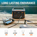 Image of Jackery Explorer 500 portable power station in outdoor camping setup on a wooden table by a lake, connected to electronic devices, front view with handle visible and endurance usage icons below.