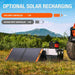 Image of Jackery Explorer 500 portable power station outdoors with optional solar panel recharging setup, angled side view showing connected solar panels and power unit on grassy terrain with mountains in the background.