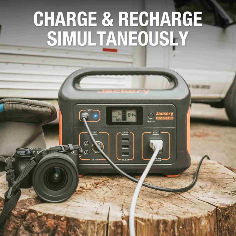 Image of Jackery Explorer 500 portable power station charging and powering devices simultaneously, front view on a wooden surface with cables connected and camera beside it in an outdoor setting.